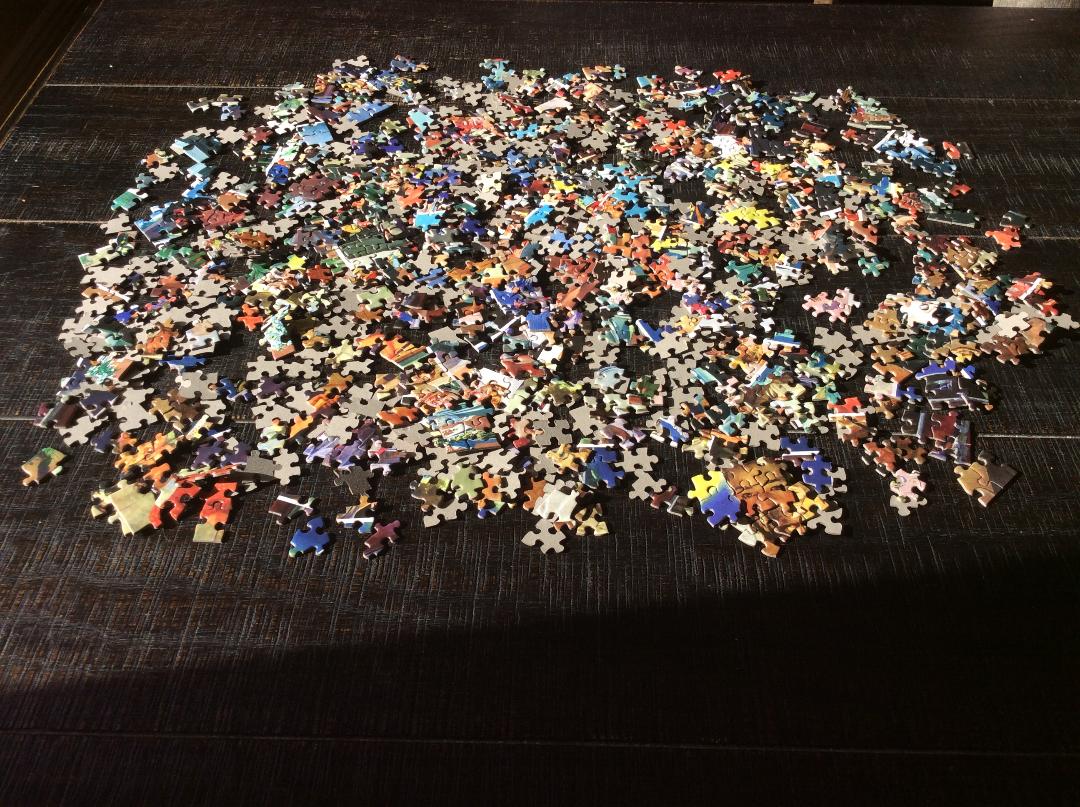 A large pile of multicolored jigsaw puzzle pieces, like scattered signs, is spread out on a dark wooden surface. The pieces, a mixture of edge and middle ones, remain unassembled. Bright sunlight illuminates the scene.