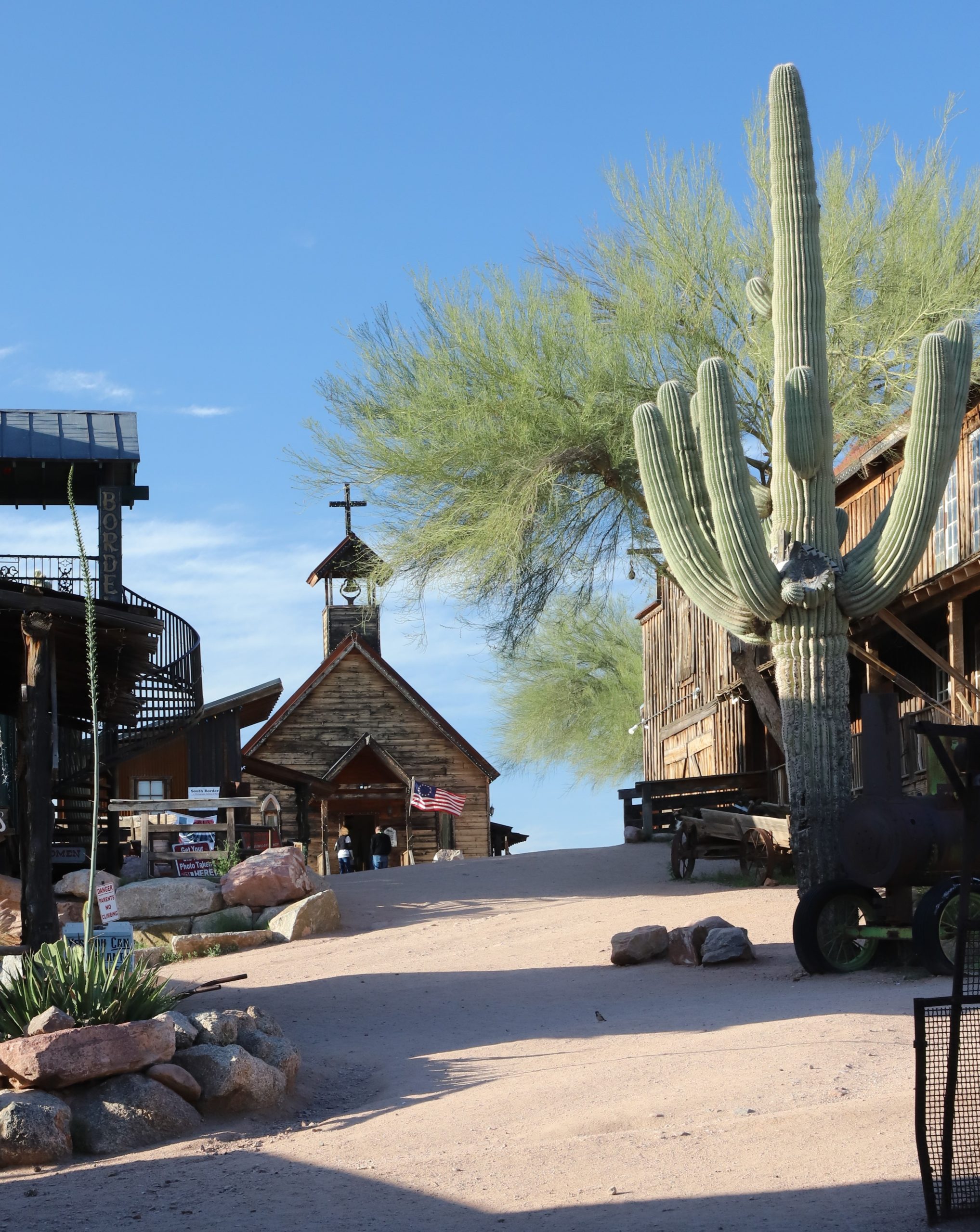 A picturesque desert scene features an old Western-style church with a cross on its roof in the background. In the foreground, a large cactus stands tall among several rustic wooden structures. The sky is clear and blue, with rocks scattered across the sandy ground.