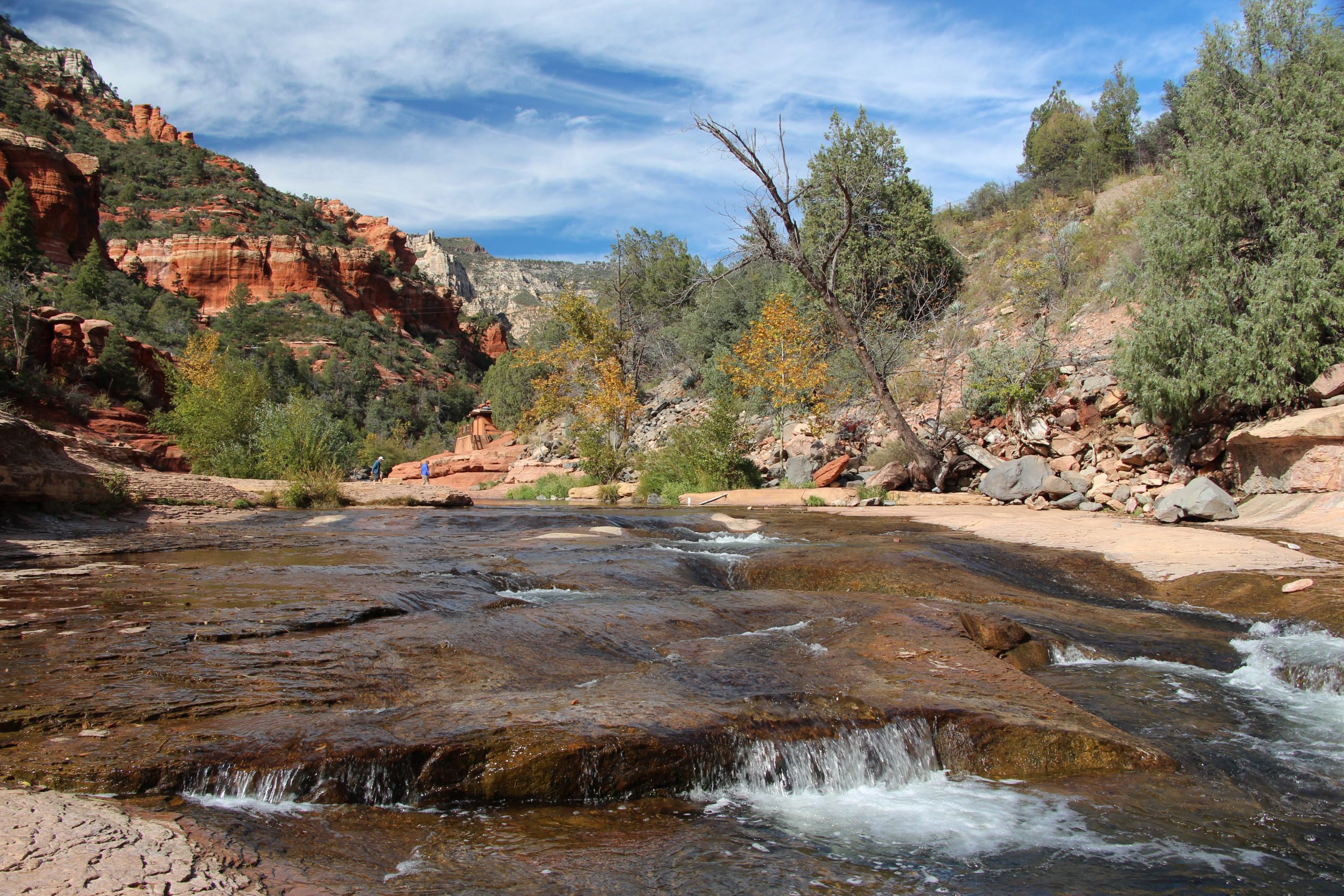A shallow stream flows over a rocky bed with small waterfalls, bordered by trees and shrubs. The background features red rock formations and a partly cloudy blue sky, creating a picturesque scene that looks like it could be straight out of a series of nature pictures.