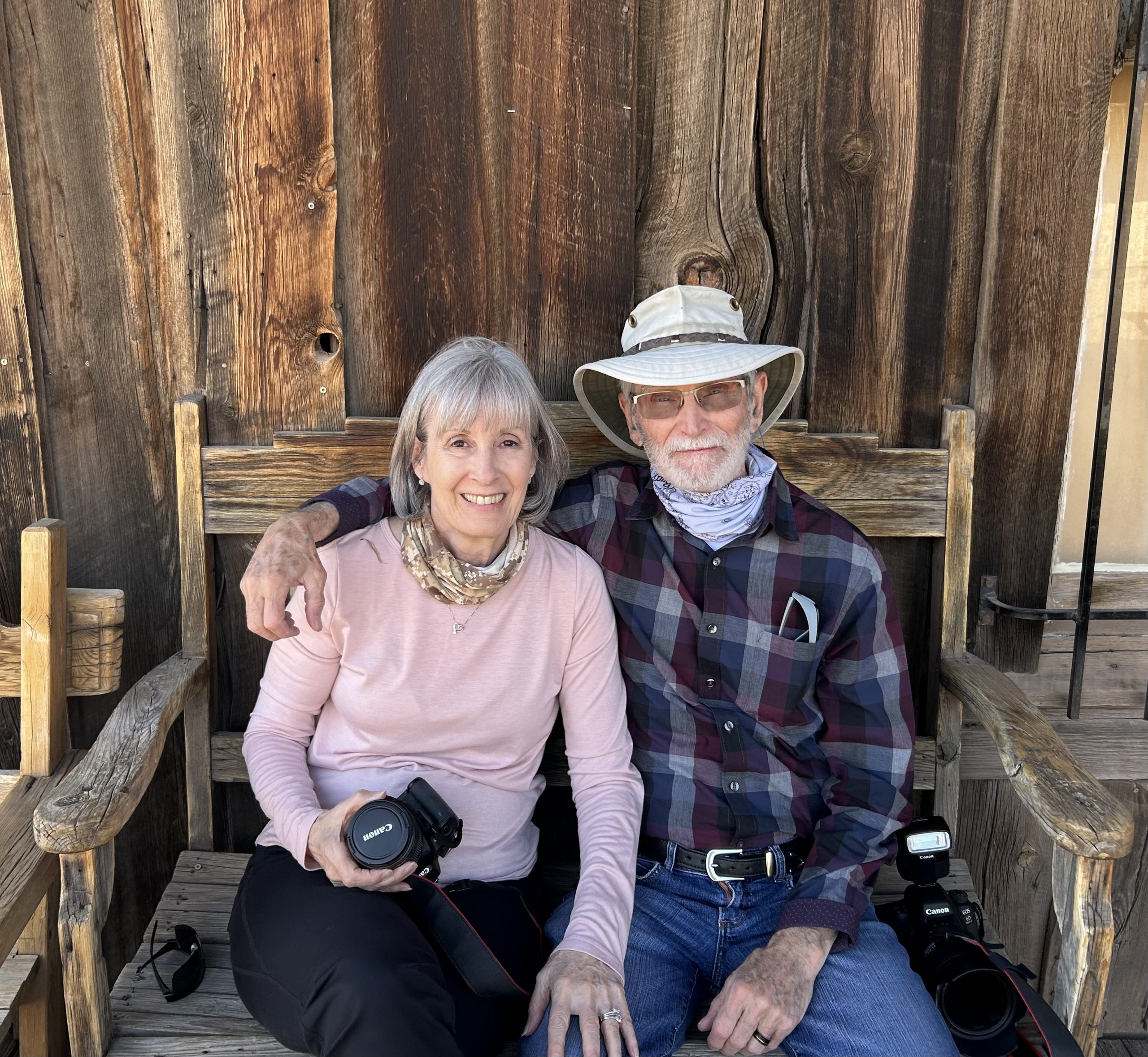 A man and woman are sitting and smiling on a wooden bench outside a rustic wooden building. The woman holds a camera, and the man wears a hat and bandana, with his arm around her shoulders. Both appear relaxed and content, dressed casually.