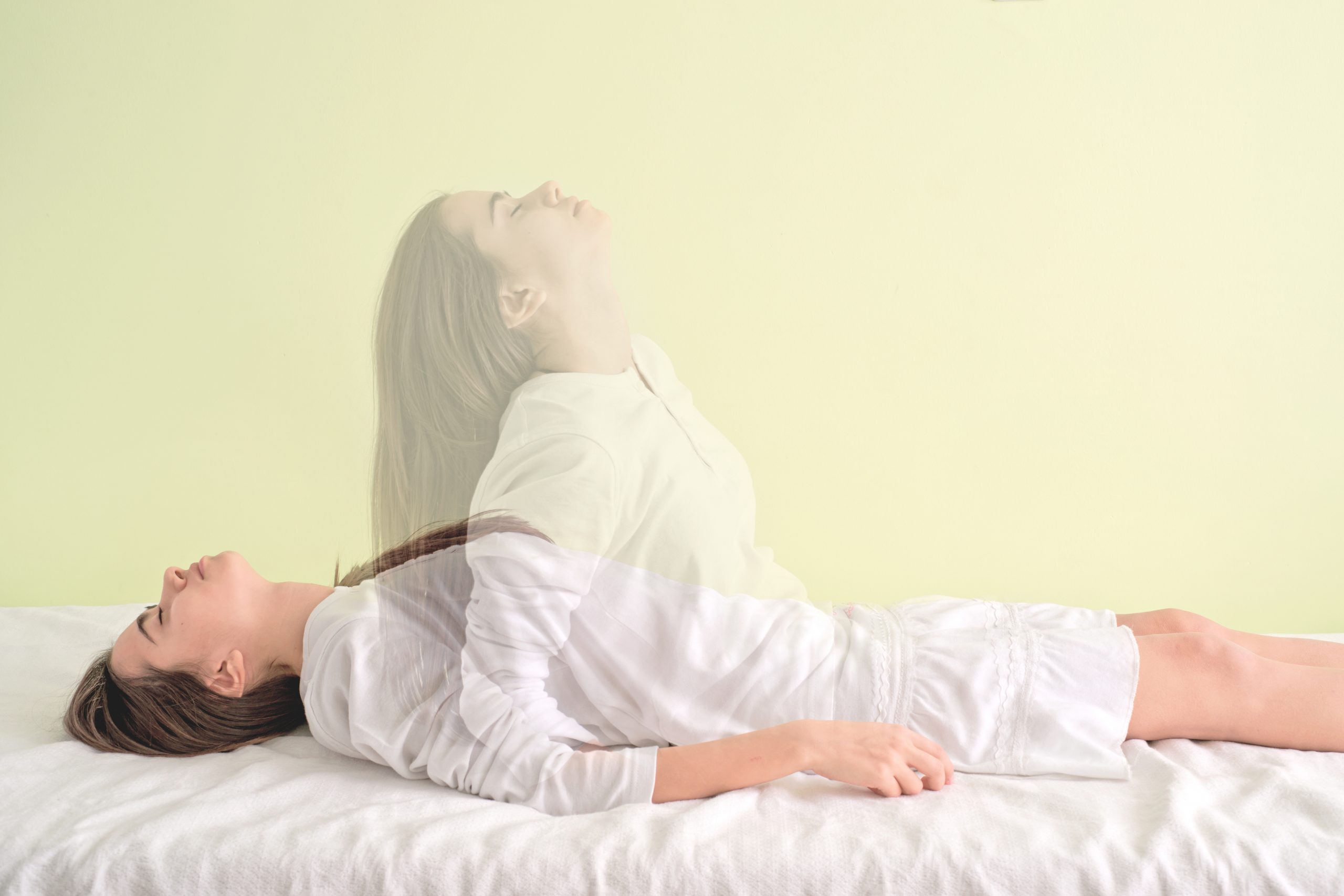 A woman lies on her back on a white surface against a light green background. Another translucent image of her spirit rises above, giving an impression of her soul leaving her body. She is wearing a white outfit.